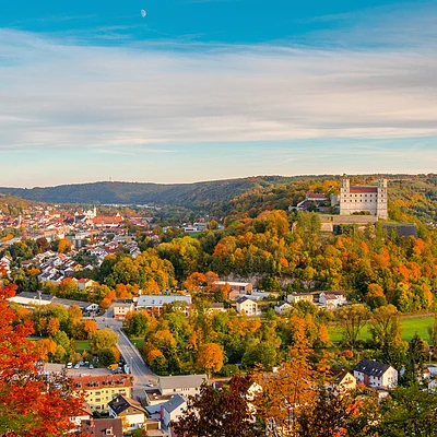 Herbstliche Landschaft mit Schloss auf Hügel, bunten Bäumen und Dorf unter blauem Himmel.