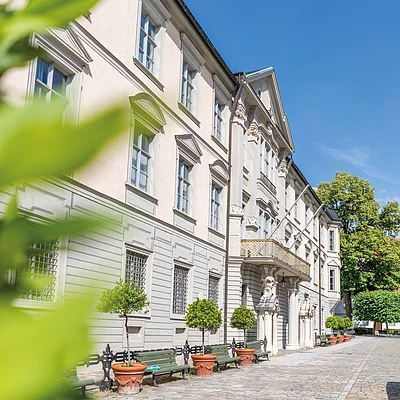Fassade eines historischen Gebäudes mit Bänken und bepflanzten Töpfen auf gepflastertem Platz bei klarem Himmel.