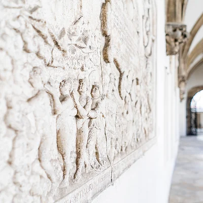 Relief mit Figuren an einer Wand in einem Gang mit gotischen Gewölben und Bogenfenstern.