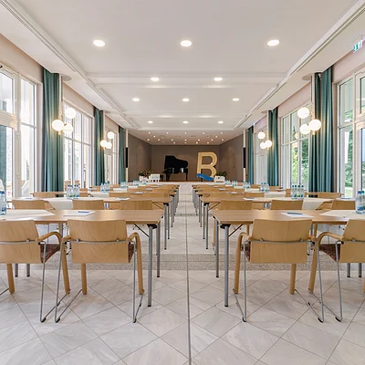 Konferenzraum mit Reihen von Tischen und Stühlen, Wasserflaschen und Notizblöcken, Bühne mit Klavier und großem Buchstaben B.