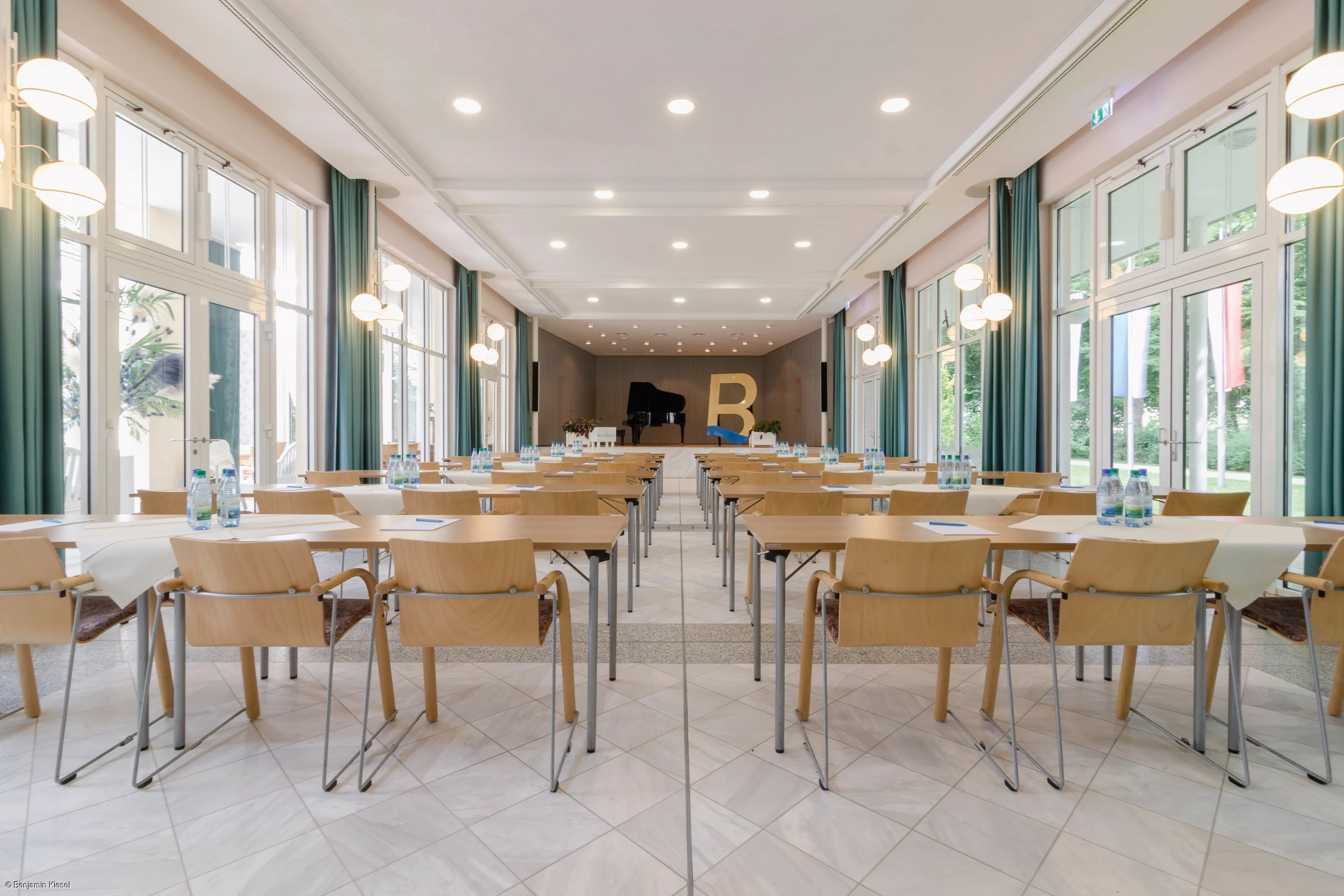 Konferenzraum mit Reihen von Tischen und Stühlen, Wasserflaschen und Notizblöcken, Bühne mit Klavier und großem Buchstaben B.