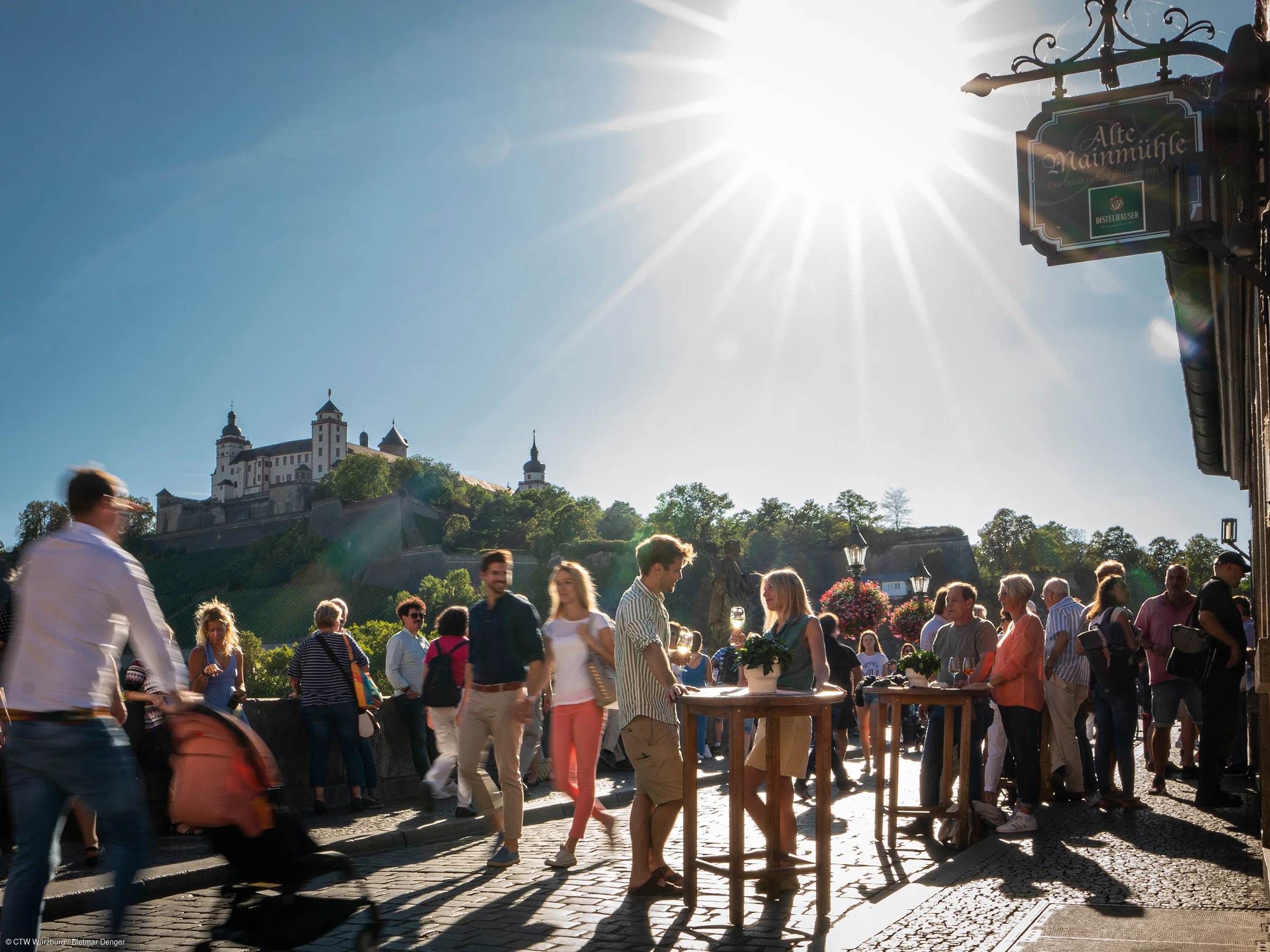 Menschen stehen und gehen auf gepflasterter Brücke bei sonnigem Himmel vor Schloss auf Hügel, Schild Alte Mainmühle sichtbar