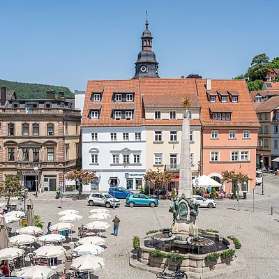 Marktplatz mit Springbrunnen, Sitzbereichen mit Sonnenschirmen und historischen Gebäuden bei klarem Himmel.