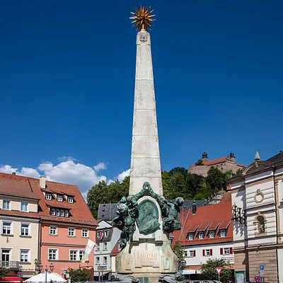 Obelisk mit bronzenem Relief und goldener Sternspitze auf gepflastertem Platz mit Blumen und Häusern im Hintergrund