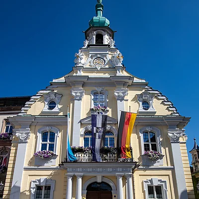 Historisches gelbes Rathaus mit Säulen, Blumenschmuck und drei Fahnen, zwei Personen vor dem Eingang bei blauem Himmel