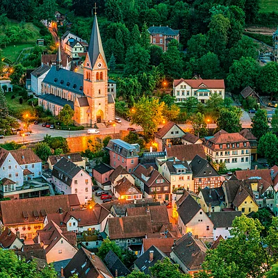 Blick auf ein Dorf mit Kirche, Häusern und Bäumen bei Abenddämmerung mit Straßenbeleuchtung.