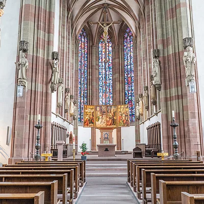 Innenraum einer Kirche mit Holzbänken, Altar, Kreuz und bunten Glasfenstern im Hintergrund