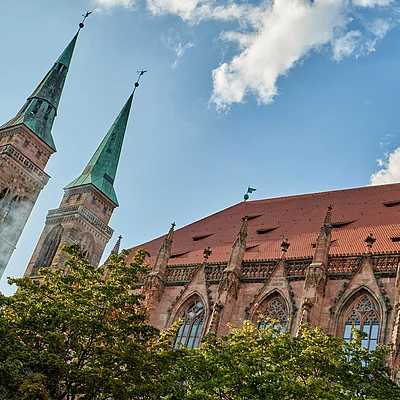 Gotische Kirche mit zwei hohen Türmen und grünem Dach vor blauem Himmel und Bäumen im Vordergrund