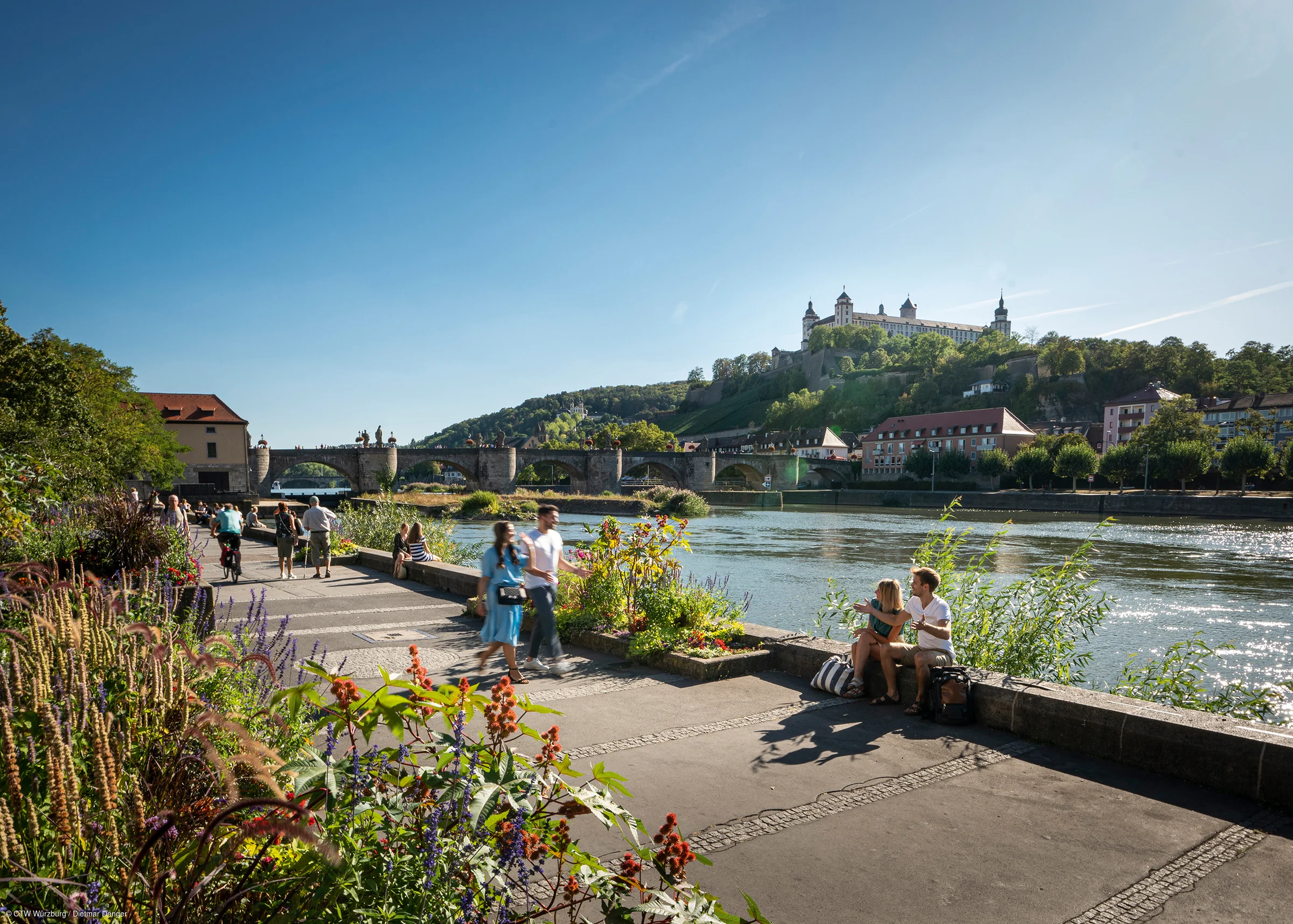 Menschen spazieren und sitzen an einem Flussufer mit Blumen, im Hintergrund Brücke und Burg auf Hügel.