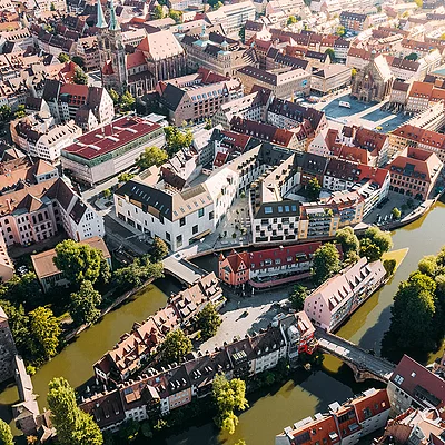 Luftaufnahme einer Altstadt mit Flussarmen, Brücken, roten Dächern und grünen Bäumen bei Sonnenschein.