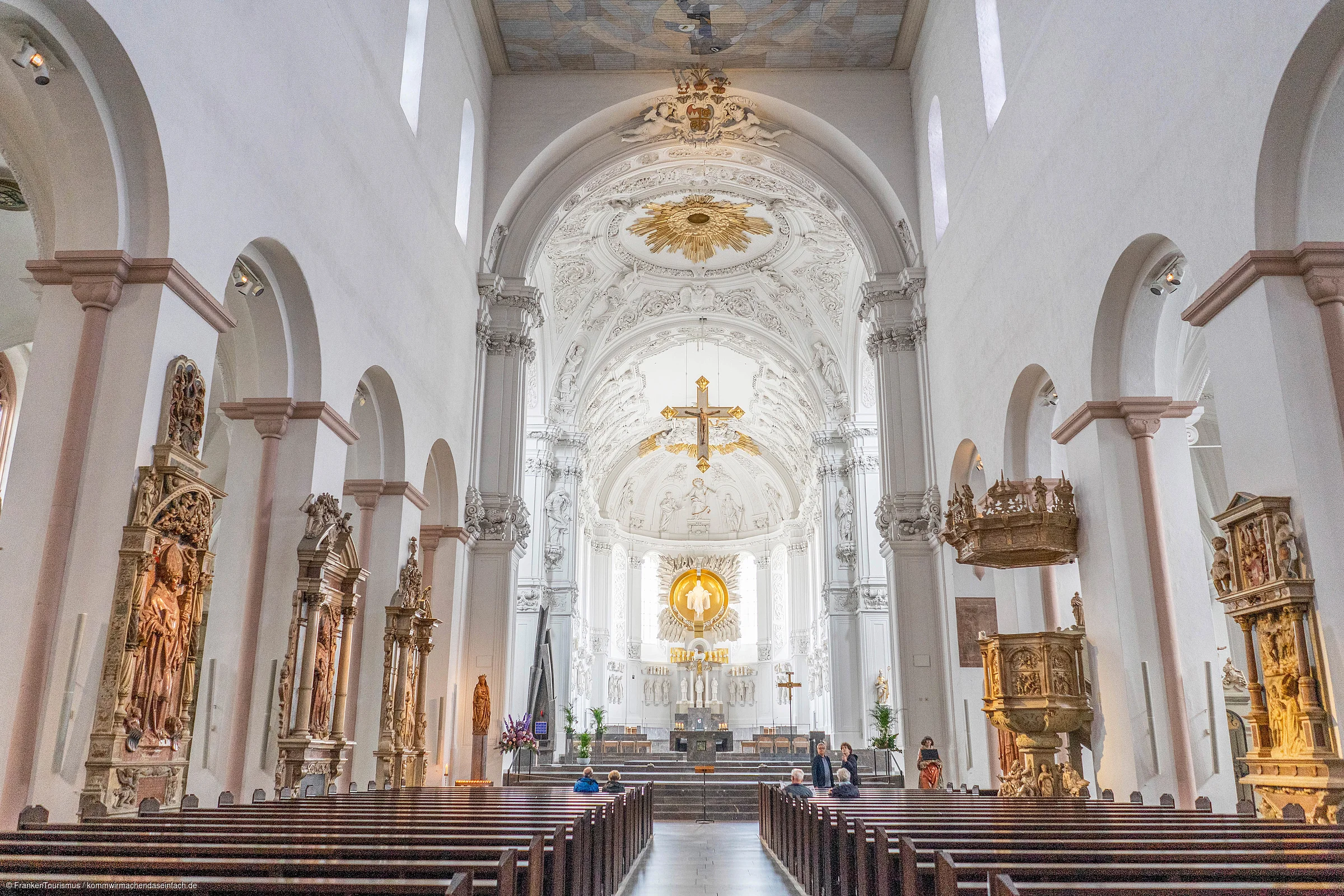 Innenraum einer Kirche mit weißen Wänden, verzierten Altären, Holzbänken und einem goldenen Kreuz über dem Altar.