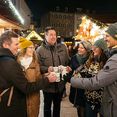 Sechs Personen stoßen mit Tassen auf einem beleuchteten Weihnachtsmarkt im Freien an.