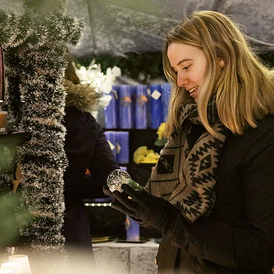 Frau mit Winterjacke und Schal betrachtet eine Schneekugel auf einem Weihnachtsmarkt im Winter.