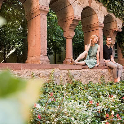 Zwei Personen sitzen an einer historischen Sandsteinarkade, davor ein Beet mit Blumen und Efeu.