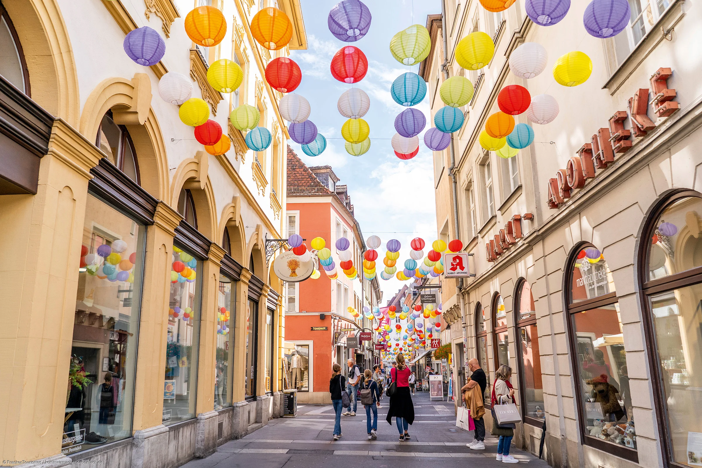 Fußgängerzone mit bunten Lampions über der Straße und Menschen, umgeben von Geschäften.