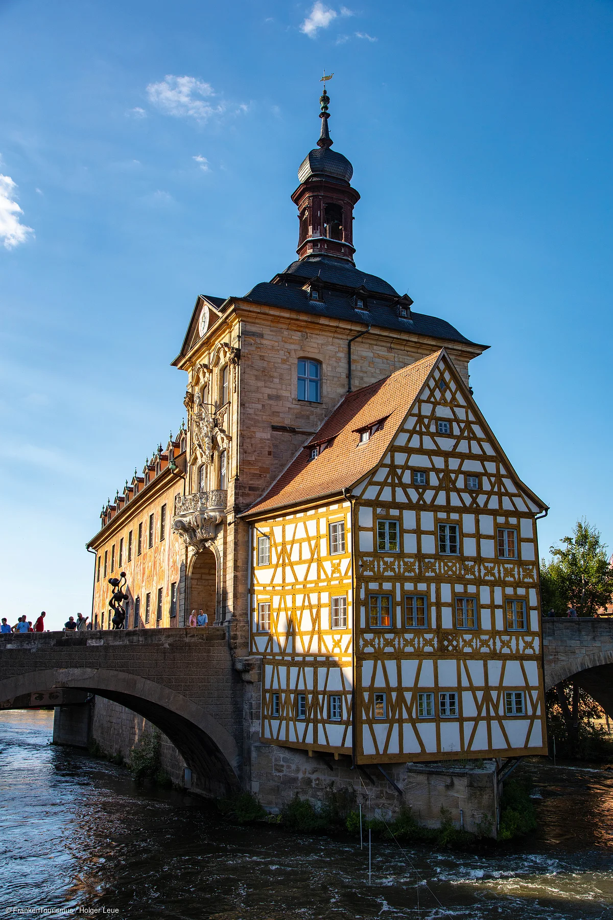 Historisches Fachwerkhaus mit Turm auf einer Steinbrücke über einen Fluss bei blauem Himmel.