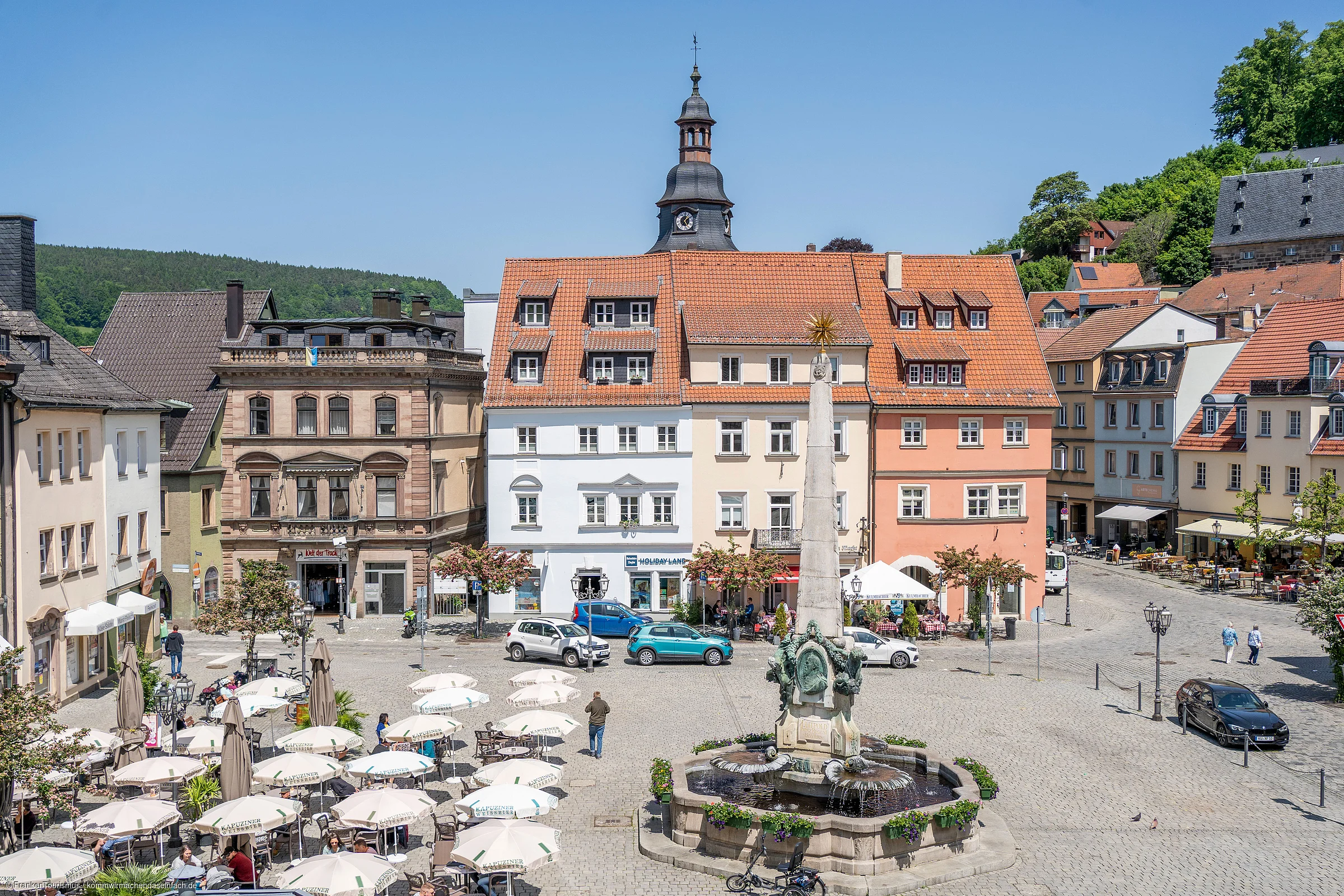 Marktplatz mit Springbrunnen, Sitzbereichen mit Sonnenschirmen und historischen Gebäuden bei klarem Himmel.