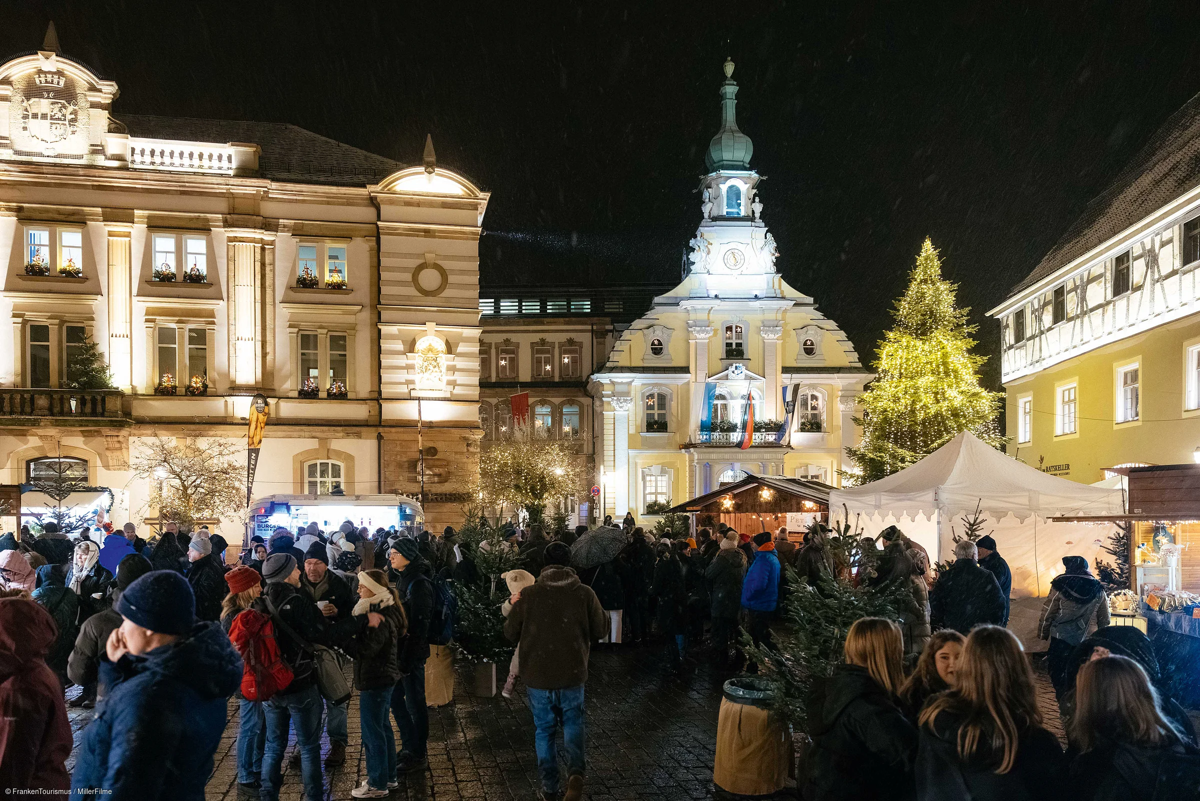 Weihnachtsmarkt mit Menschen, beleuchteten Gebäuden und Weihnachtsbaum bei Nacht in einer Stadt.