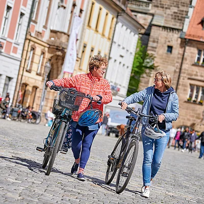 Zwei Frauen schieben Fahrräder auf einem gepflasterten Platz vor historischen Gebäuden.
