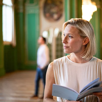 Frau mit kurzem blonden Haar hält ein geöffnetes Buch in einem großen Raum mit grünen Wänden.