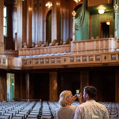 Zwei Personen fotografieren in einem großen Saal mit vielen Sitzreihen und einer hölzernen Königsloge.