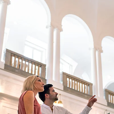 Zwei Personen betrachten und zeigen auf eine Ausstellungstafel in einem hellen Gebäude mit hohen Bögen.