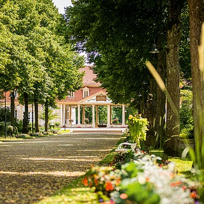 Baumallee mit Blumenrabatten und Sitzbank führt zu einem historischen Gebäude mit Säulenvorbau.