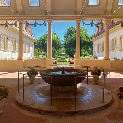 Innenhof mit Springbrunnen, Säulen und Blumendekoration vor einem Garten mit Bäumen und blauem Himmel