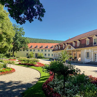 Garten mit Blumenbeeten, Bäumen und zwei Gebäuden mit roten Dächern unter blauem Himmel.