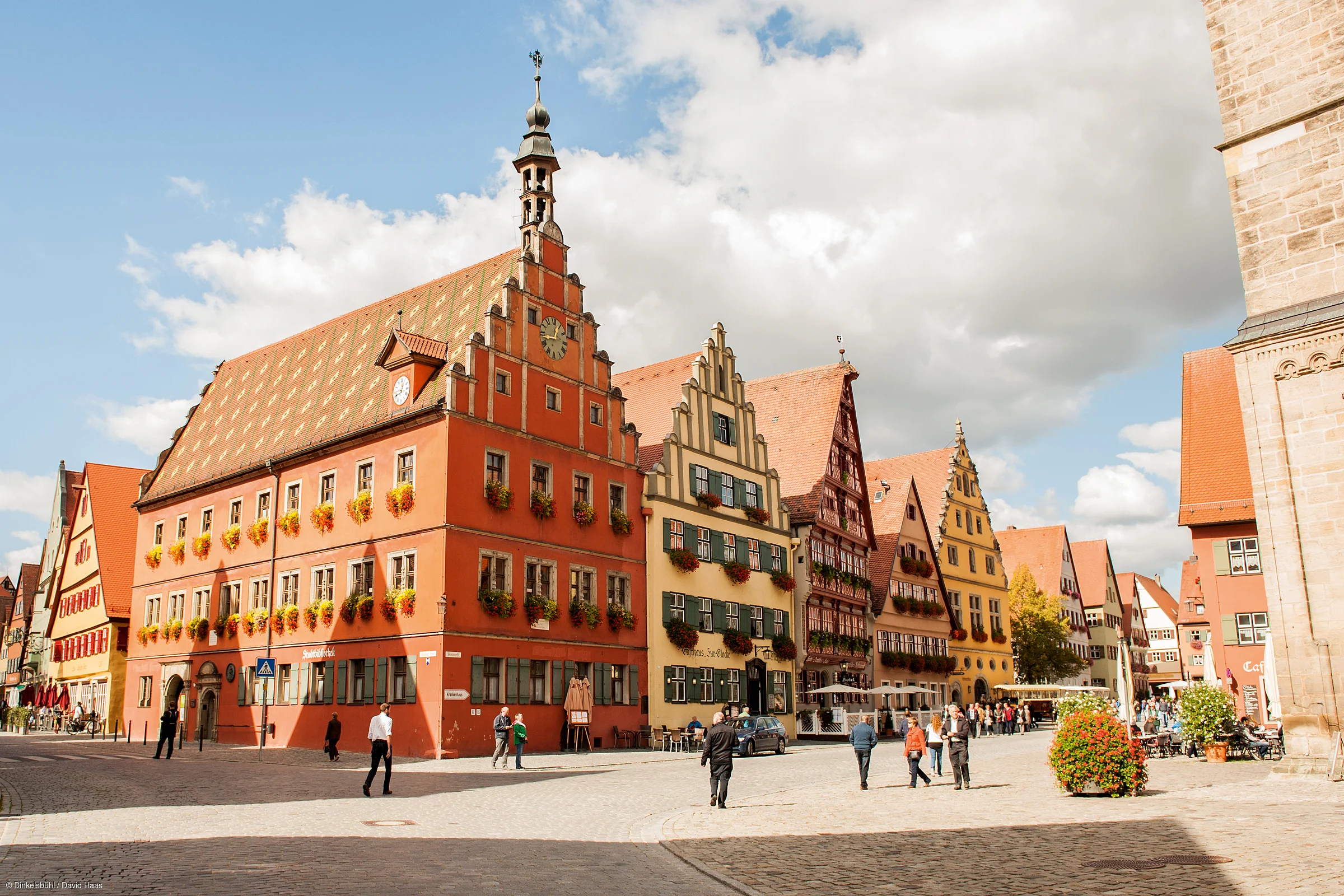 Historische Fachwerkhäuser mit Blumenschmuck an Fenstern und Menschen auf gepflastertem Platz bei Tageslicht.