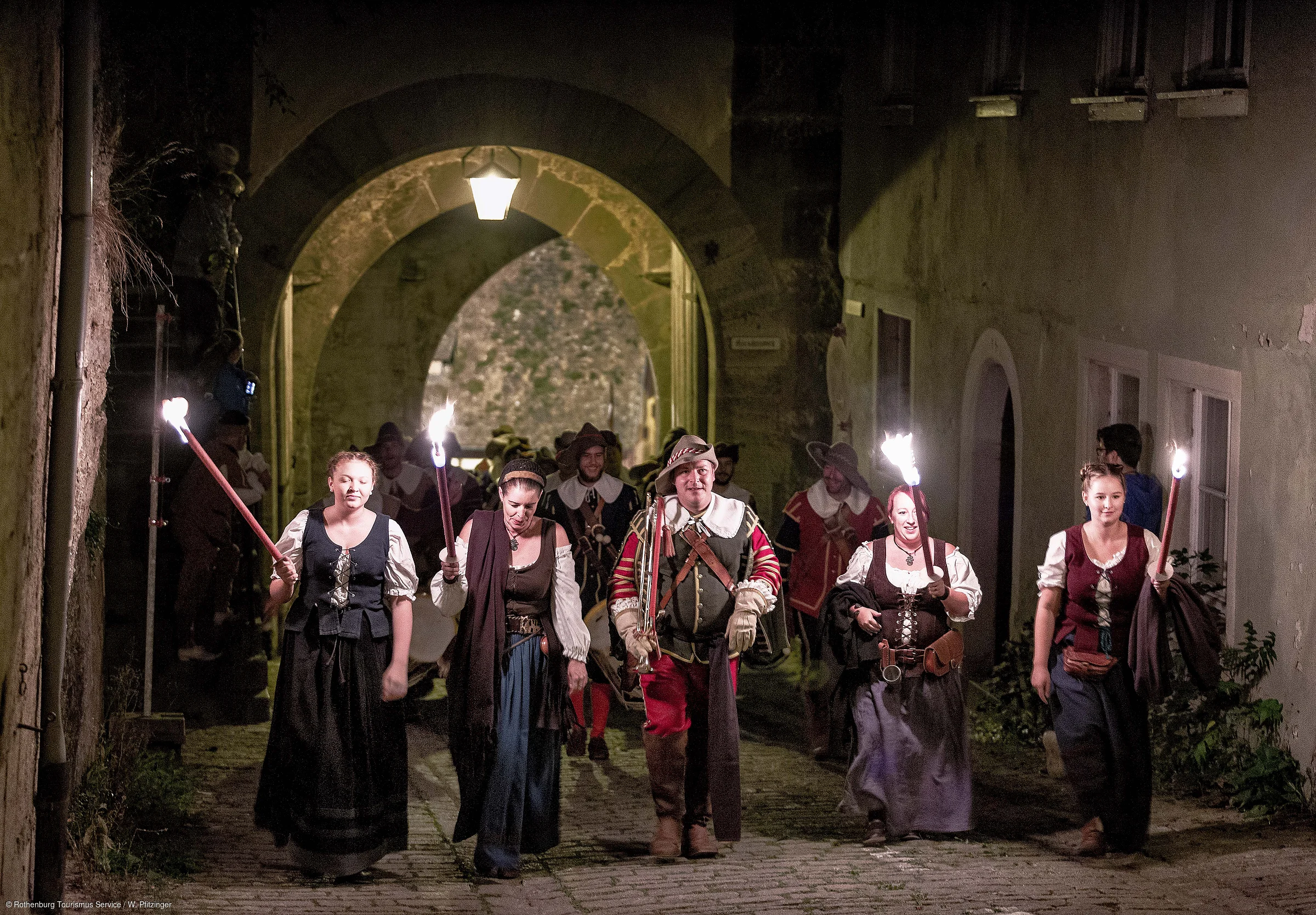 Gruppe in historischen Kostümen mit Fackeln geht bei Nacht durch einen beleuchteten Torbogen auf Kopfsteinpflaster.