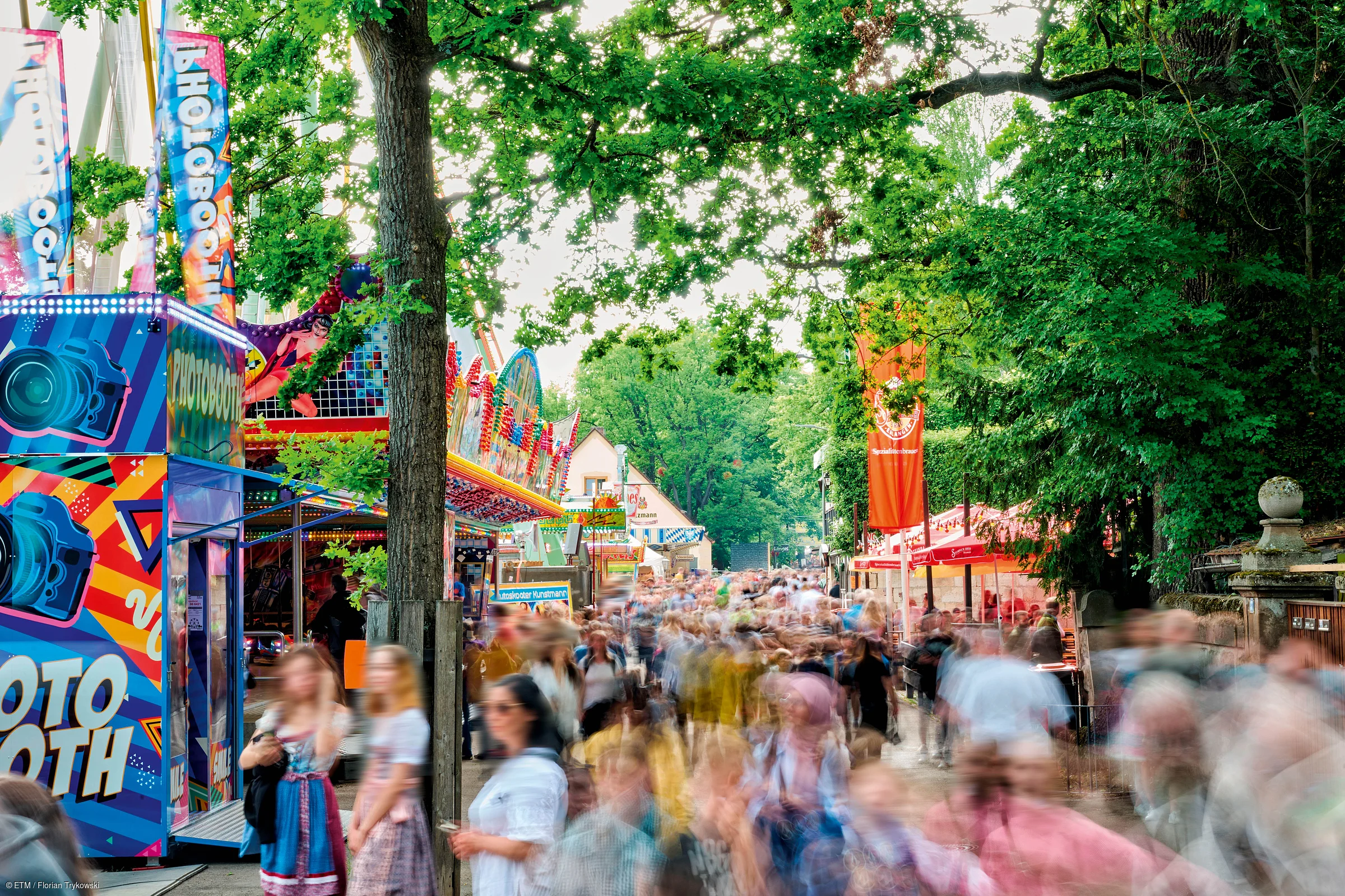 Menschenmenge auf einem Fest mit Fahrgeschäften und Bäumen an einem sonnigen Tag