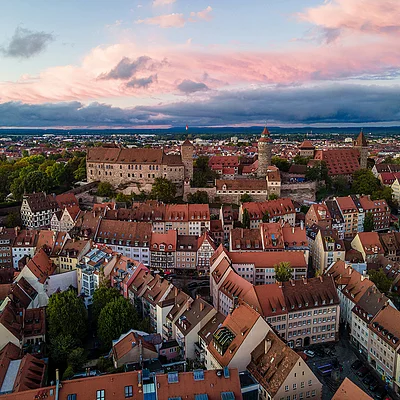 Luftaufnahme einer Stadt mit roten Dächern und historischen Türmen bei Sonnenuntergang und bewölktem Himmel.