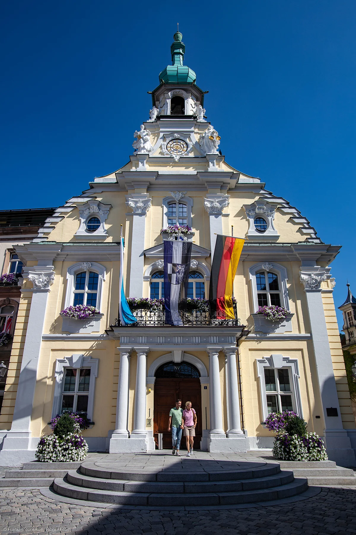 Historisches gelbes Rathaus mit Säulen, Blumenschmuck und drei Fahnen, zwei Personen vor dem Eingang bei blauem Himmel