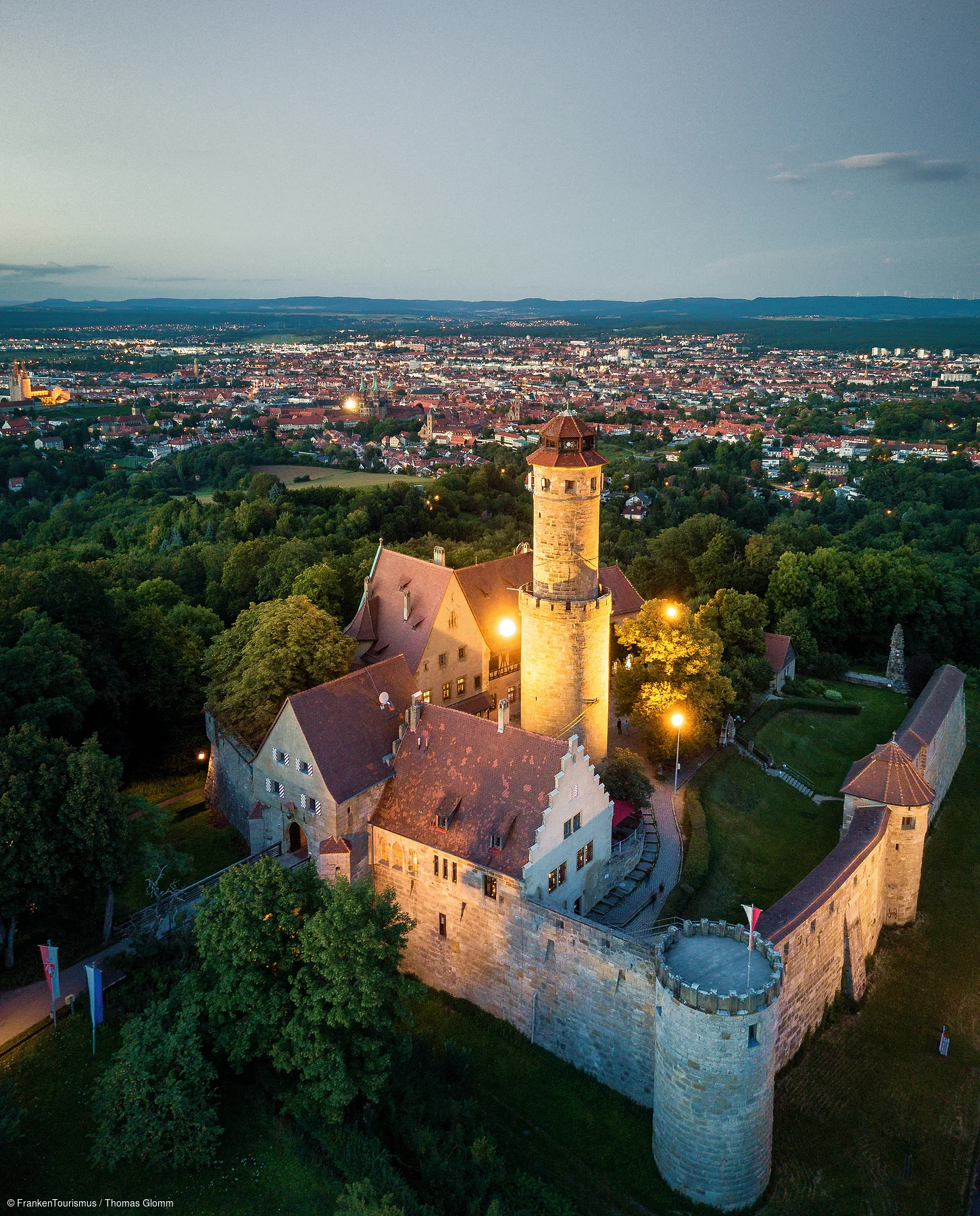 Luftaufnahme einer beleuchteten Burg mit Stadt und Wald im Hintergrund bei Abenddämmerung.