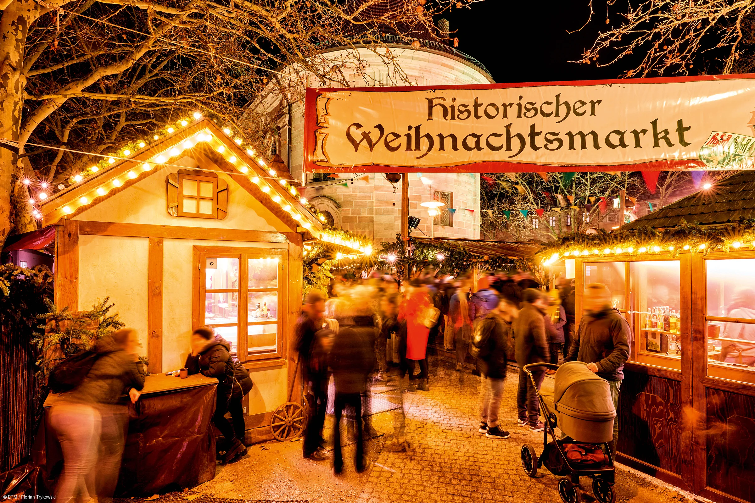 Menschen auf historischem Weihnachtsmarkt mit beleuchteten Buden und Banner bei Nacht.