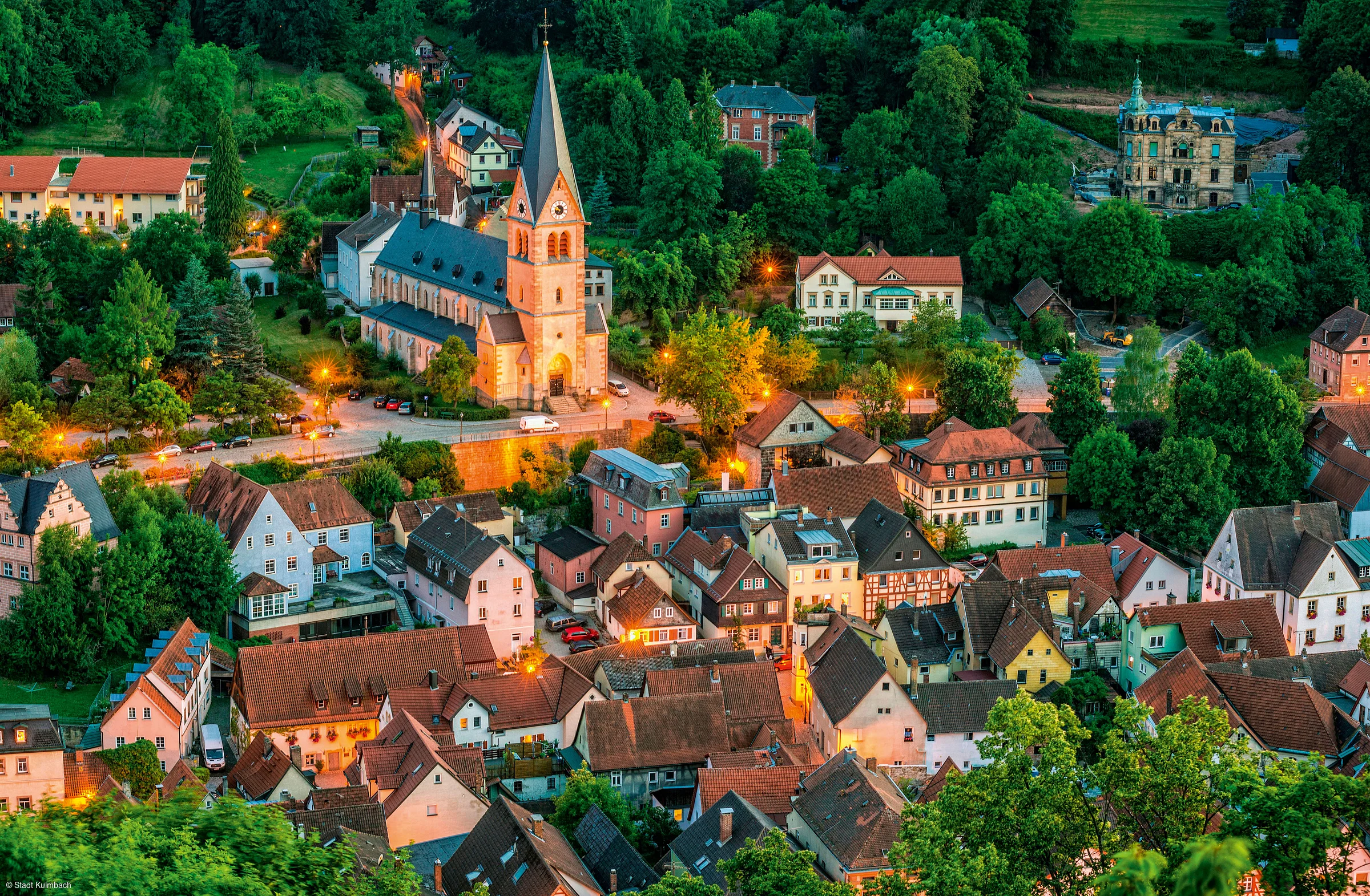 Blick auf ein Dorf mit Kirche, Häusern und Bäumen bei Abenddämmerung mit Straßenbeleuchtung.