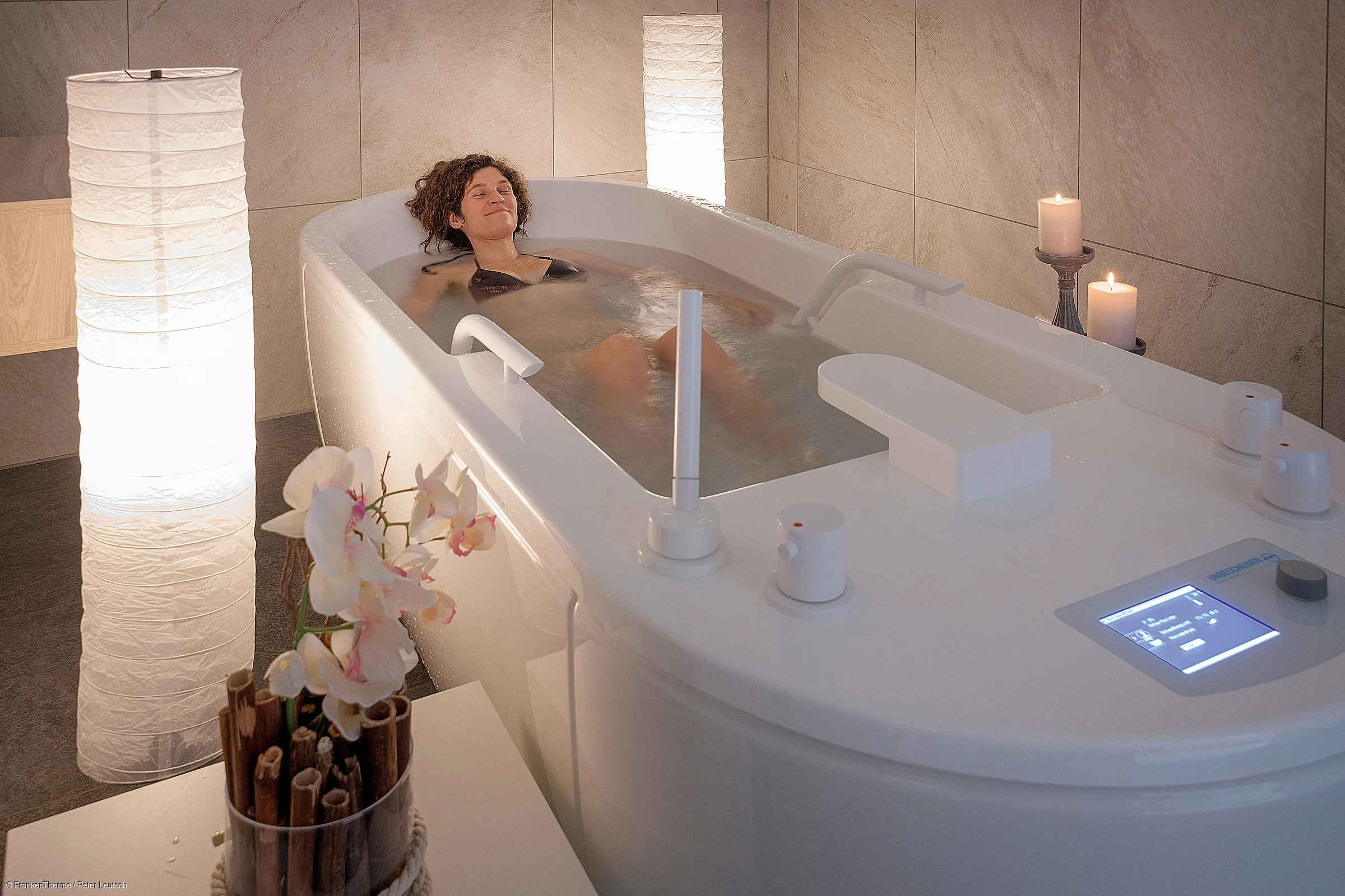 Frau entspannt in moderner Badewanne mit Wasser, Kerzen, Orchidee und sanfter Beleuchtung.