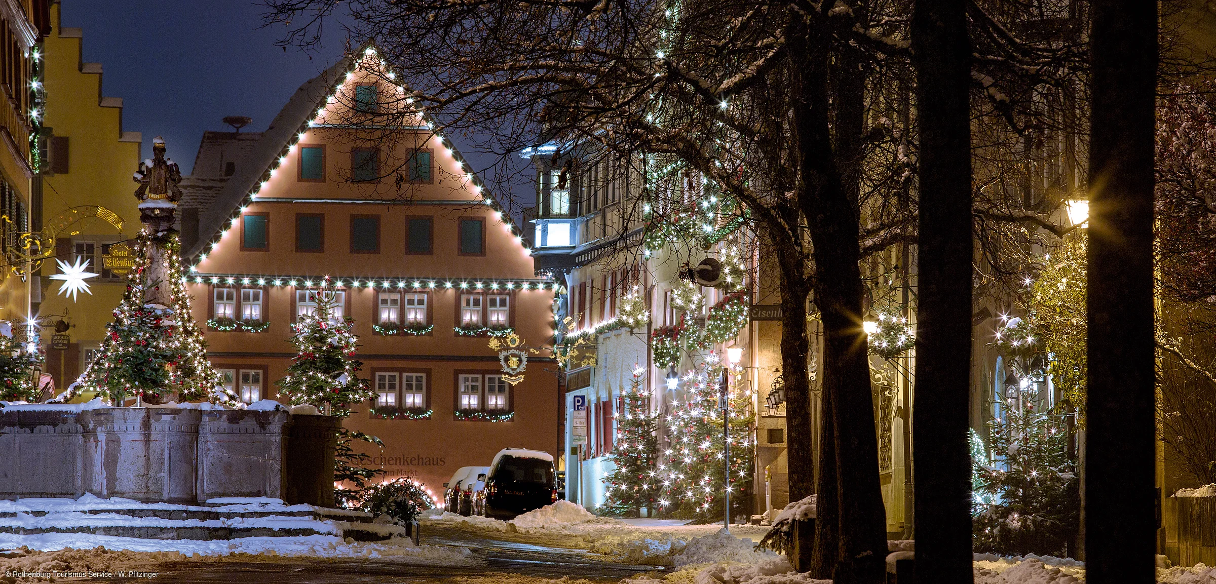 Weihnachtlich beleuchtete Fachwerkhäuser und geschmückte Tannenbäume an einem verschneiten Abend in einer Altstadt