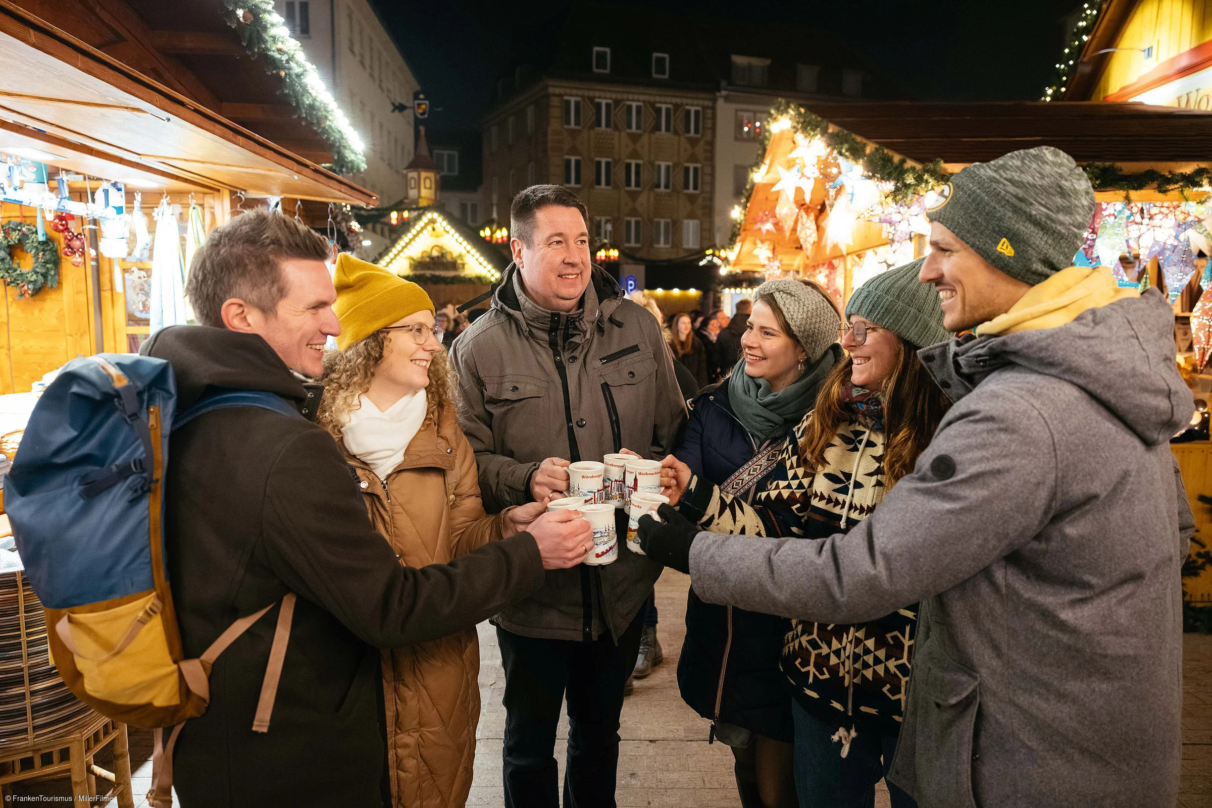 Sechs Personen stoßen mit Tassen auf einem beleuchteten Weihnachtsmarkt im Freien an.