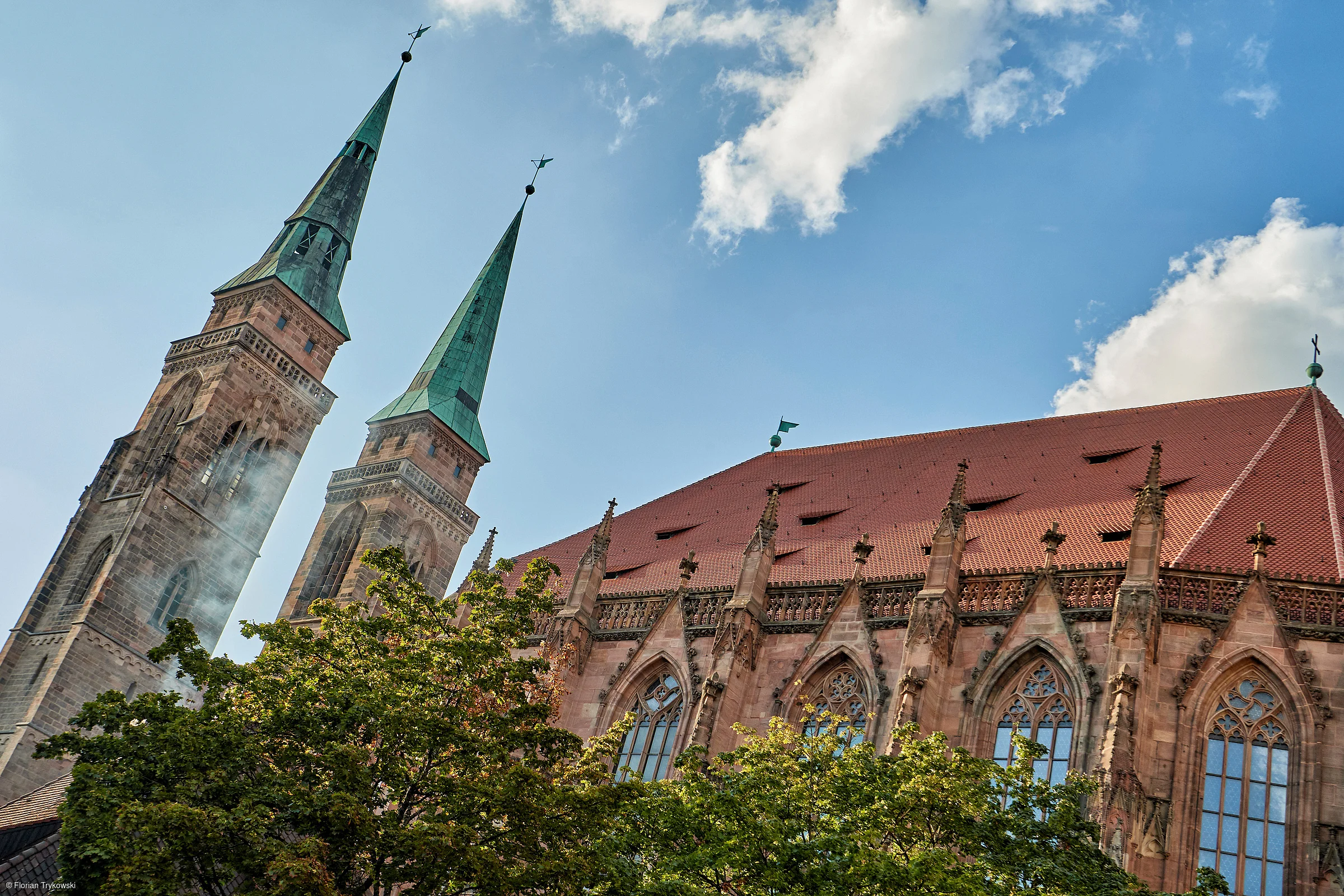 Gotische Kirche mit zwei hohen Türmen und grünem Dach vor blauem Himmel und Bäumen im Vordergrund
