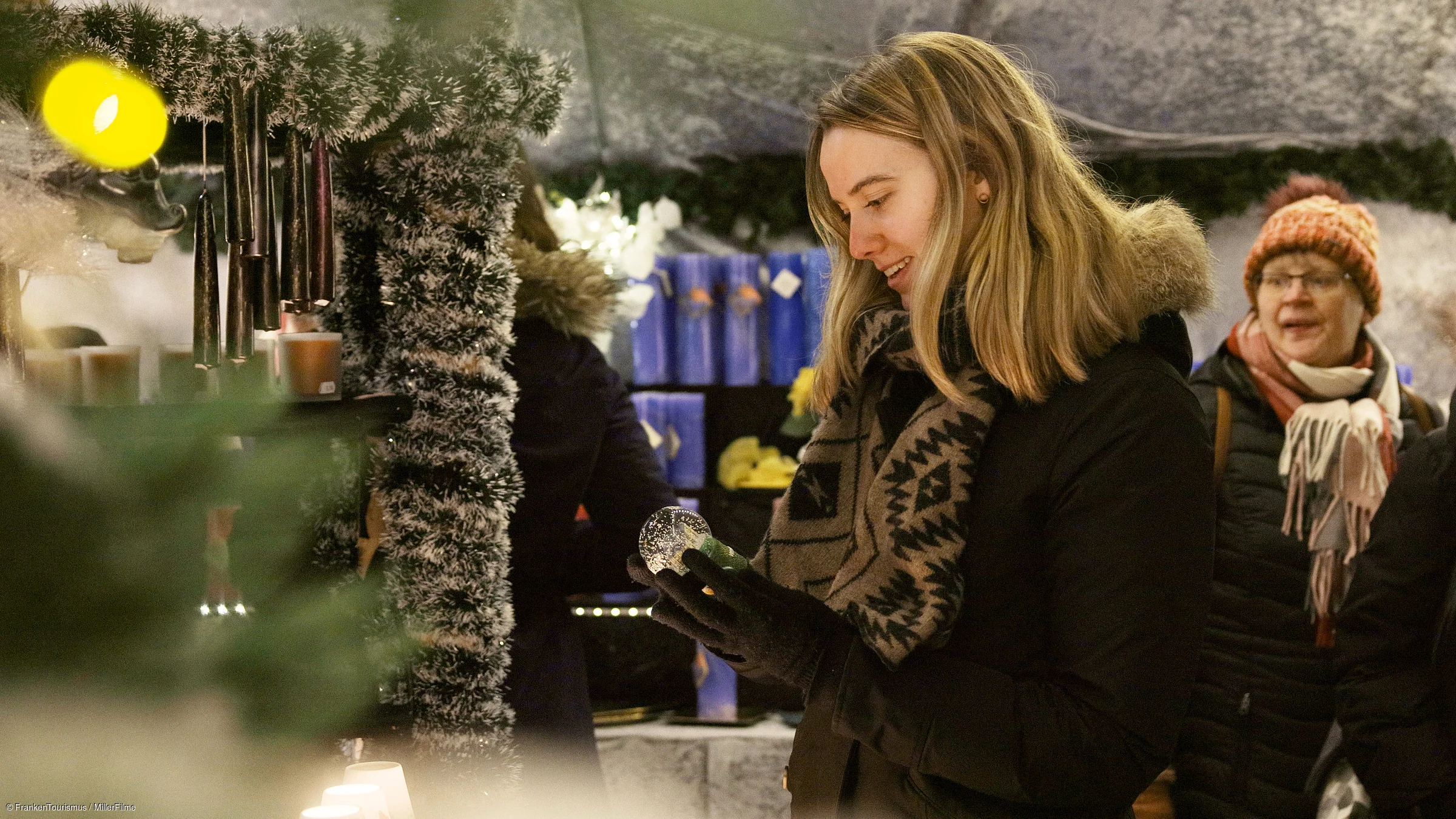 Frau mit Winterjacke und Schal betrachtet eine Schneekugel auf einem Weihnachtsmarkt im Winter.