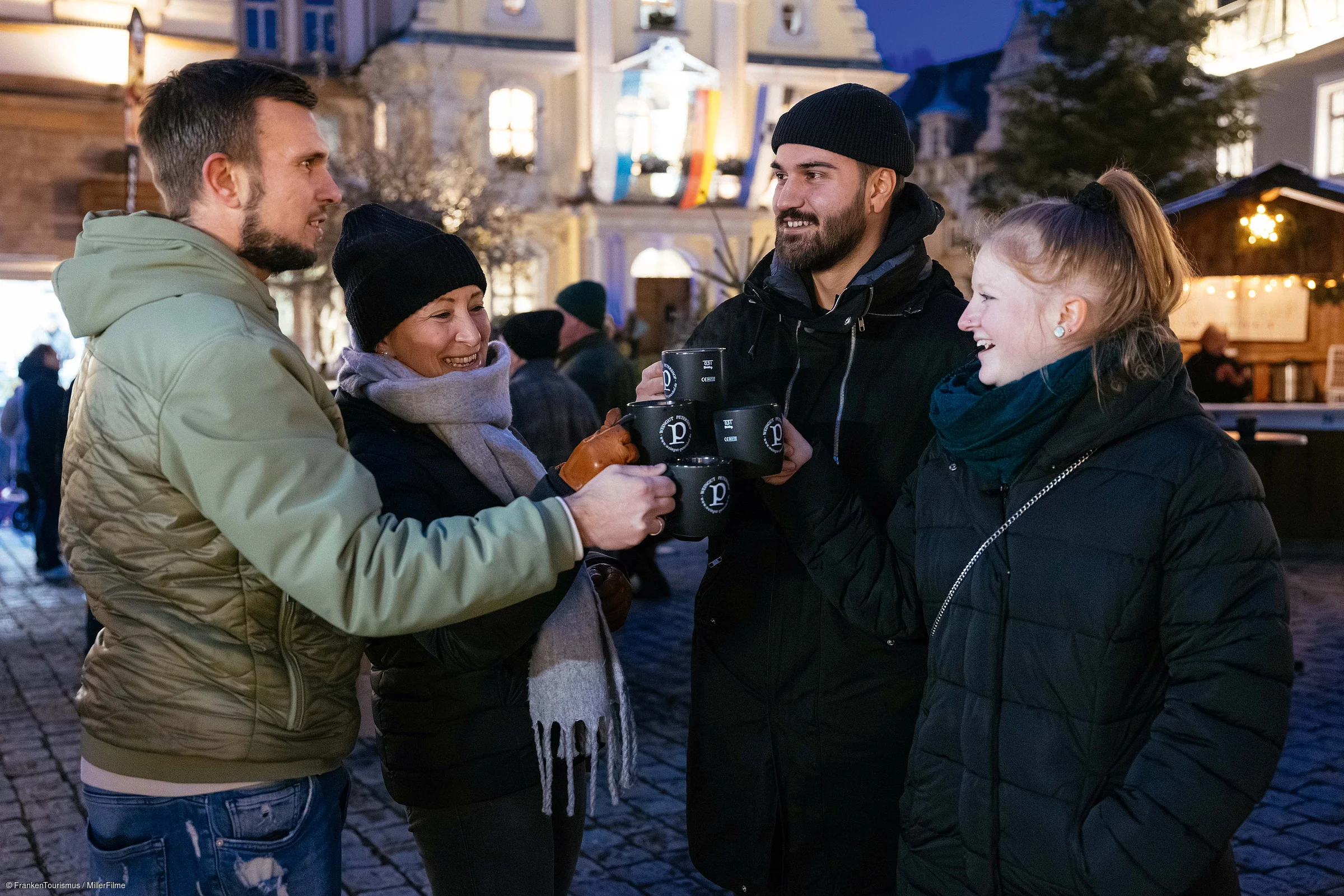 Vier Personen stoßen mit schwarzen Bechern bei einem abendlichen Treffen im Freien an.
