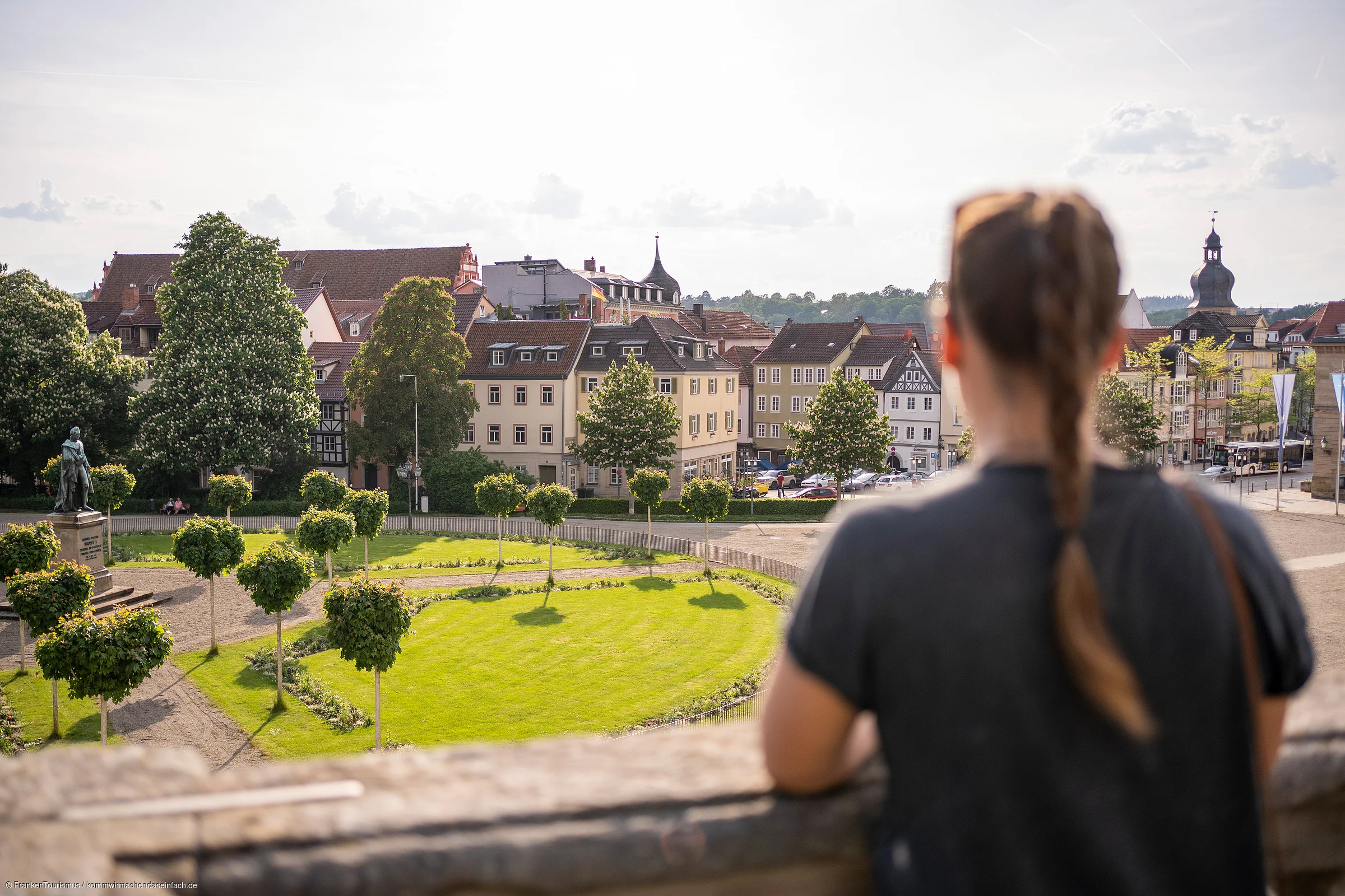 Frau mit geflochtener Frisur blickt auf Park mit Bäumen und historische Gebäude im Hintergrund.