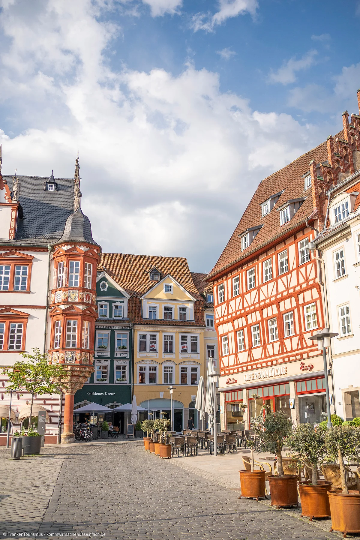 Historische Fachwerkhäuser und Außengastronomie an gepflastertem Platz unter bewölktem Himmel.