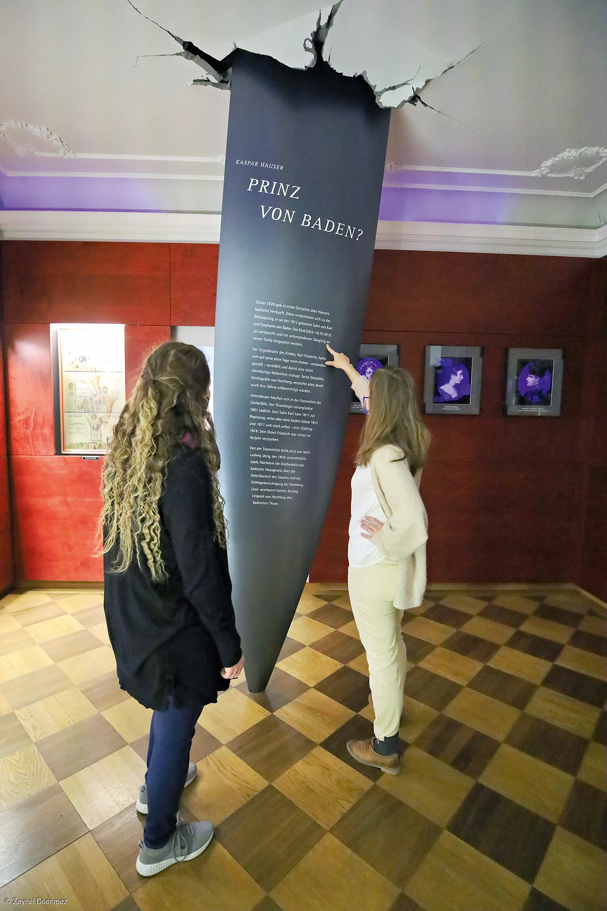 Zwei Frauen betrachten eine Museumsausstellung mit Texttafel, die aus der Decke zu brechen scheint.