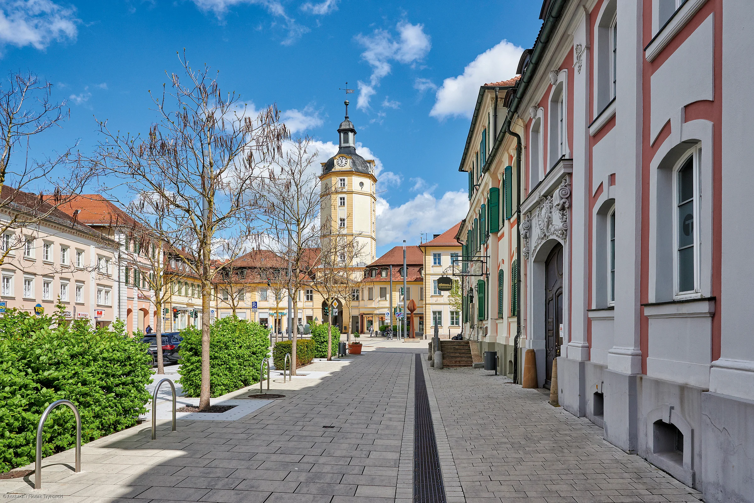 Blick auf eine gepflasterte Straße mit kahlen Bäumen, Gebäuden und einem Uhrturm im Hintergrund bei blauem Himmel.