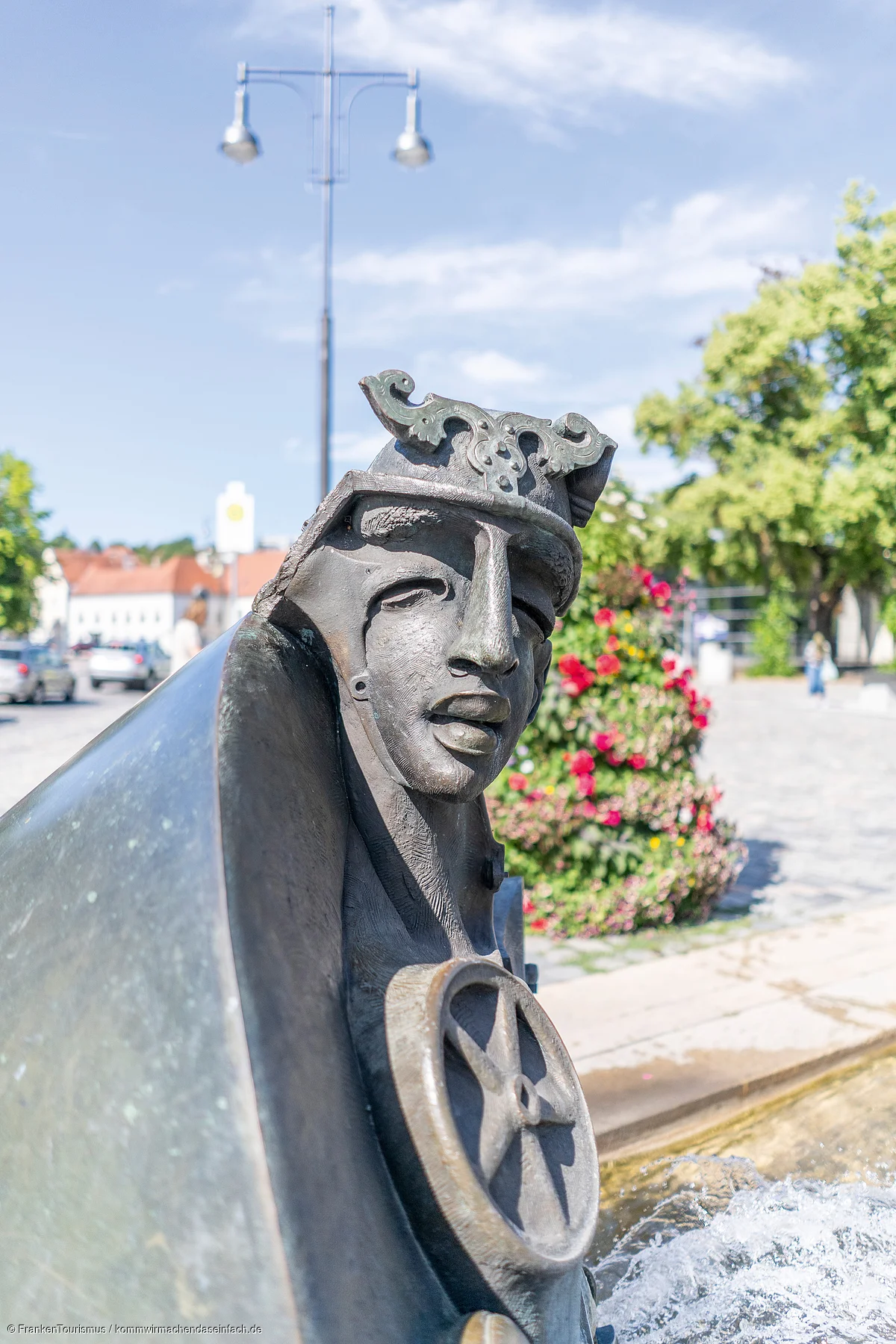 Bronzefigur mit Helm und Rad an einem Brunnen in einer Stadt mit Blumen und Bäumen im Hintergrund
