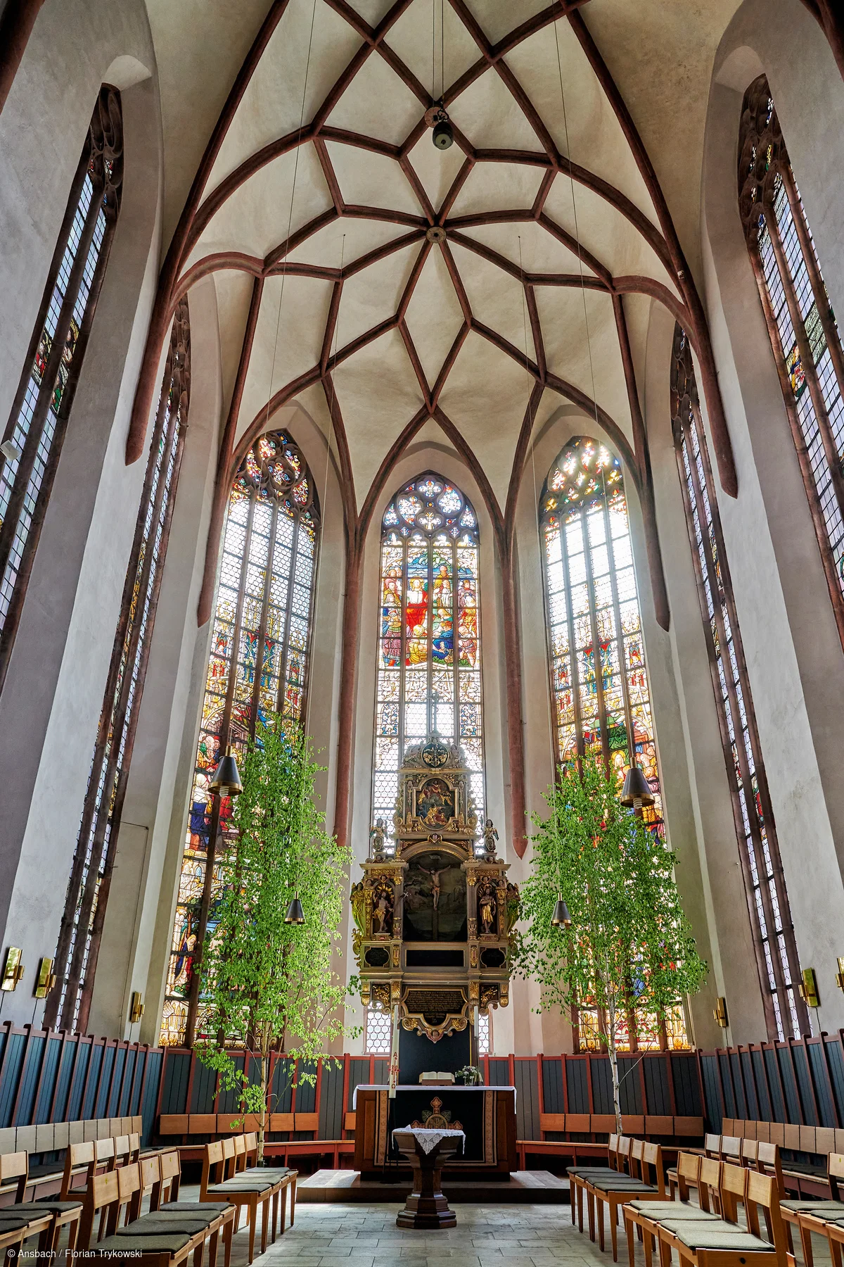Innenraum einer Kirche mit hohen Buntglasfenstern, Holzbänken und einem Altar mit zwei grünen Bäumen.