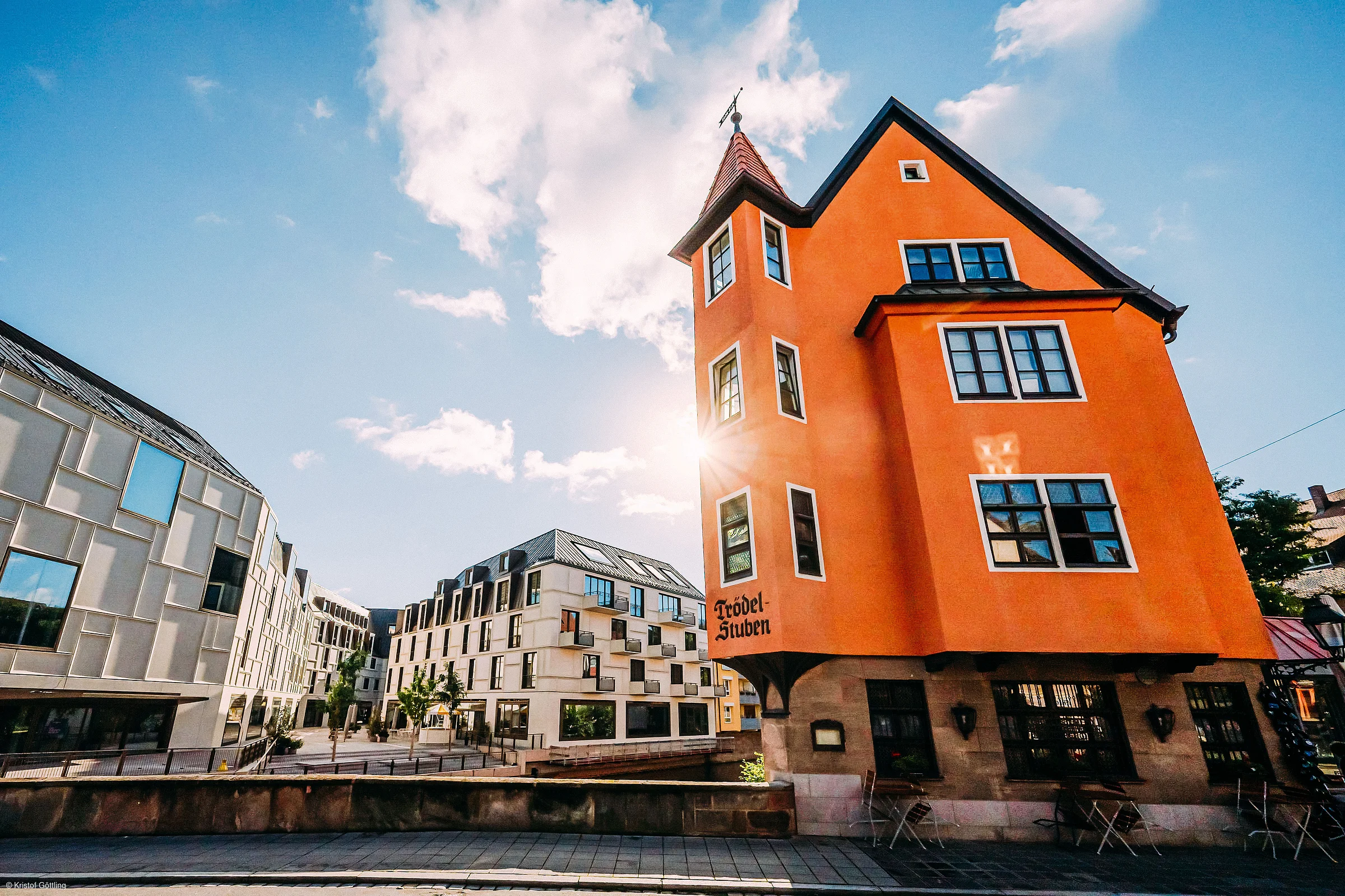 Oranger Altbau mit Turm neben modernen weißen Gebäuden bei sonnigem Himmel und Wolken.
